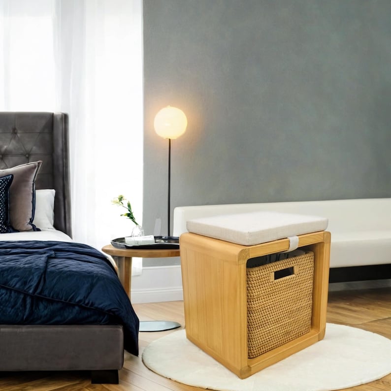 May include: A light wood storage bench with a white cushion and a woven basket inside. The bench is in a bedroom setting with a bed and a side table.