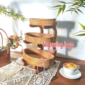 May include: A three-tiered serving stand with woven rattan baskets and a wooden frame. The stand features oval-shaped baskets and is suitable for displaying food or decorative items. The text "Engraving logo" is visible.