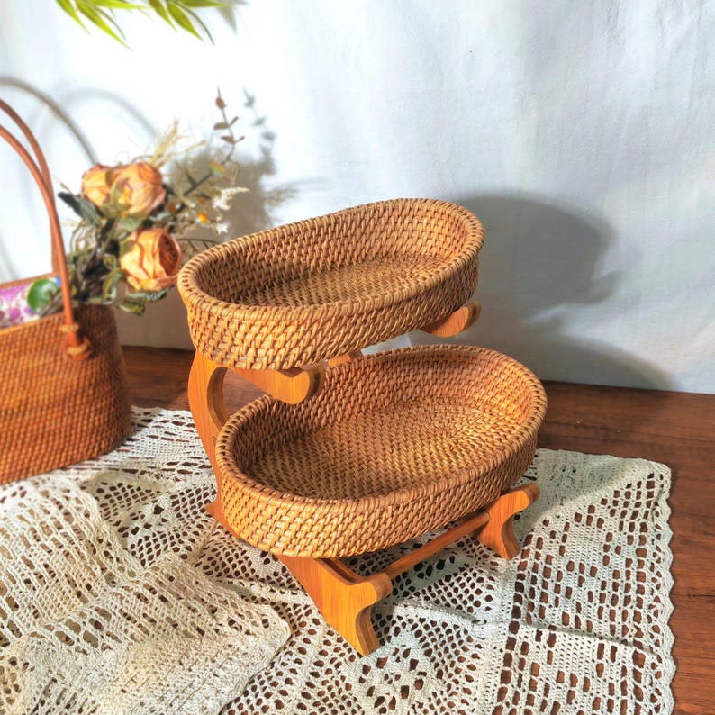 May include: Two-tiered woven wicker serving stand with a natural wood finish. The stand is made of natural rattan and features two oval-shaped baskets.