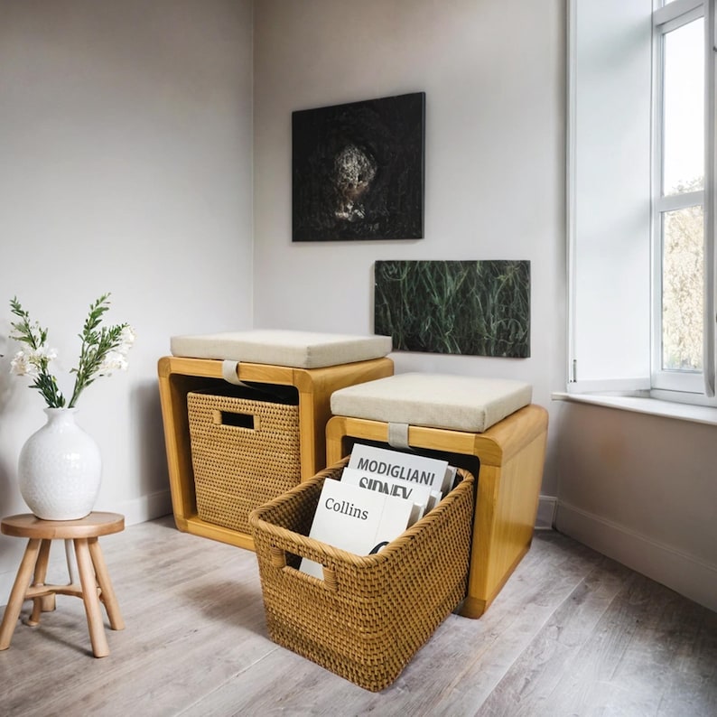 May include: Two wooden storage ottomans with beige cushions and woven baskets. The ottomans are in a room with white walls and artwork. One basket is filled with books, including "Modigliani".