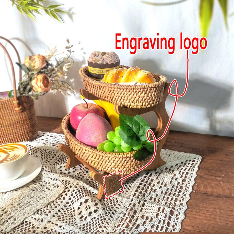 May include: Two-tiered woven wicker fruit basket with a wooden stand. The stand has an engraved logo. The basket is filled with fruit, including apples, grapes, and a peach. The top tier has a small cake and a loaf of bread.