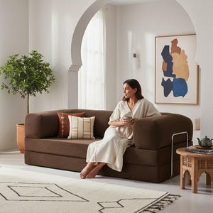 Puede incluir: Un sof&aacute; modular marr&oacute;n con cojines decorativos, una peque&ntilde;a mesa auxiliar de madera y una alfombra estampada. Una mujer est&aacute; sentada en el sof&aacute;, sosteniendo una taza. Una obra de arte abstracta enmarcada cuelga de la pared.