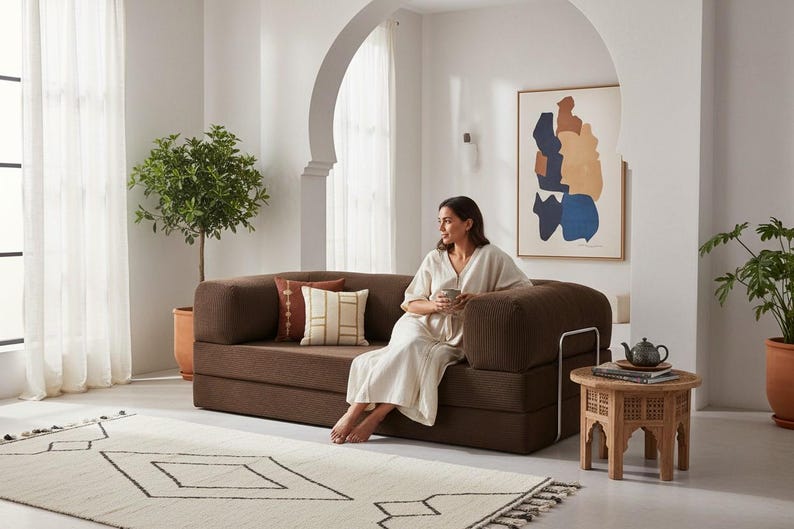 Puede incluir: Un sof&aacute; modular marr&oacute;n con cojines decorativos, una peque&ntilde;a mesa auxiliar de madera y una alfombra estampada. Una mujer est&aacute; sentada en el sof&aacute;, sosteniendo una taza. Una obra de arte abstracta enmarcada cuelga de la pared.