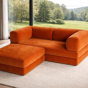 May include: An orange modular sofa and ottoman set. The sofa has a rounded arm and back design. The ottoman is square and sits flush with the sofa. The furniture is made of a soft, textured fabric. The set is placed on a light-colored rug in front of a large window.