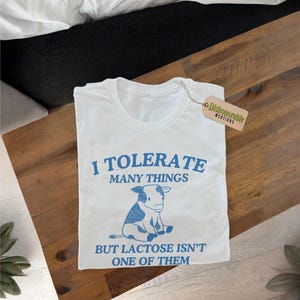 May include: White t-shirt with blue text that reads "I tolerate many things but lactose isn't one of them". A blue cartoon cow is pictured below the text.