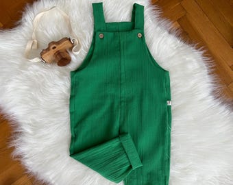 Könnte beinhalten: Eine grüne Latzhose für Kleinkinder mit Holzknöpfen und hochgekrempelten Manschetten. Eine Spielzeugkamera aus Holz mit einem cremefarbenen Riemen liegt daneben. Die Latzhose ist aus einem strukturierten Stoff gefertigt.