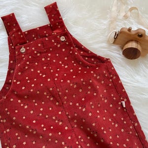 May include: Red corduroy overalls with a floral pattern. The overalls have adjustable straps and wooden buttons. A wooden toy camera with a cream-colored strap rests nearby.