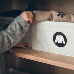 May include: A white fabric storage bin with a black circle label featuring a white silhouette of a jumper. The bin is on a wooden shelf and a child's hand is reaching into the bin to pull out a brown and white patterned shirt.