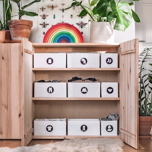 May include: A wooden cabinet with six white storage bins. The bins have different symbols on them, including a dress, a shirt, trousers, and underwear. The cabinet has a rainbow-coloured arch above it.