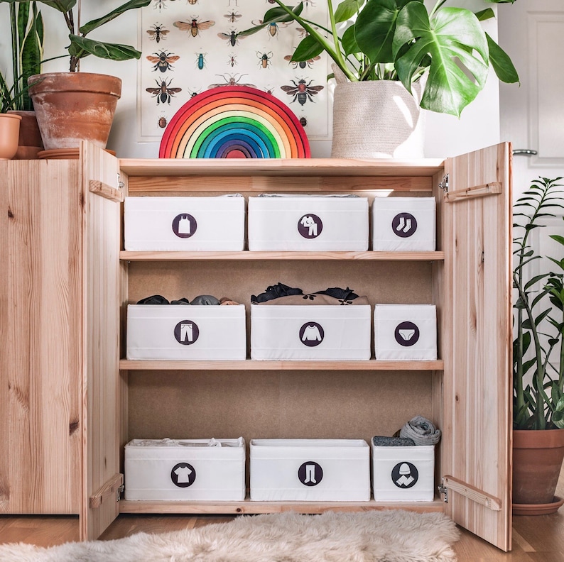 May include: A wooden cabinet with six white storage bins. The bins have different symbols on them, including a dress, a shirt, trousers, and underwear. The cabinet has a rainbow-coloured arch above it.