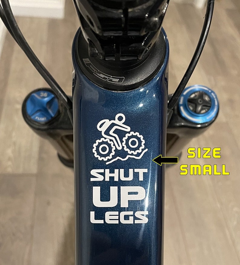 May include: A blue bicycle frame with a white sticker that says "SHUT UP LEGS" and a small graphic of a person riding a bike. The sticker also says "SIZE SMALL" in the bottom right corner.