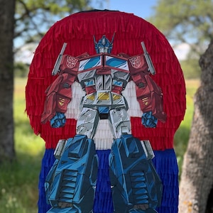 May include: An Optimus Prime piñata in red, white, and blue. The piñata depicts the Transformers character with a detailed illustration, set against a backdrop of red fringe at the top, transitioning to white and blue fringe at the bottom. It is suspended from a metal hook.