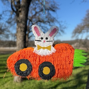 Puede incluir: Una piñata con forma de zanahoria con una cara de conejo blanco, orejas rosas y una pajarita dorada. La piñata tiene ruedas negras con centros dorados y hojas verdes. Cuelga de un hilo de pescar al aire libre.