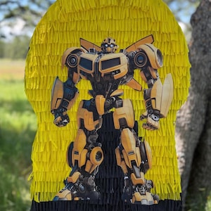 May include: A yellow piñata featuring a detailed image of the Transformers character Bumblebee. The piñata has a yellow fringe background and a black fringe base, suitable for a birthday party or themed event.