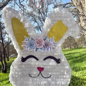 May include: White bunny piñata with gold ears, a floral crown, and a pink nose. The piñata is hanging from a string and is surrounded by green grass.