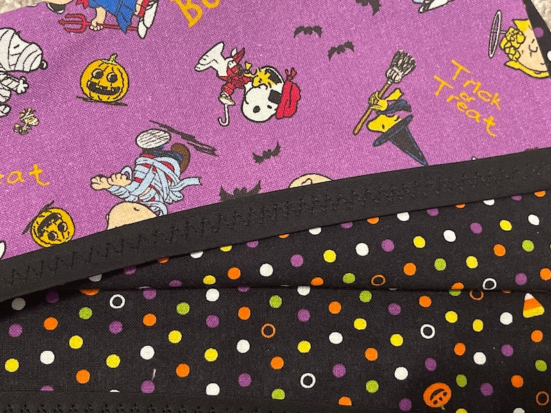May include: Close-up of Halloween-themed fabric featuring Snoopy and the Peanuts gang in costumes. The purple fabric has pumpkins and bats. Below is black fabric with white, orange, and purple polka dots. A black zigzag trim separates the fabrics.