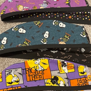 May include: Several Halloween-themed scrub caps featuring Snoopy and the Peanuts gang. The caps have black trim and are patterned with pumpkins, bats, and the words "Trick or Treat." Colors include purple, teal, and black.