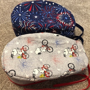 May include: Two fabric scrub caps. One is navy blue with a red, white, and blue fireworks pattern. The other is gray with a Snoopy on a bicycle and fireworks design. Both caps have ties.