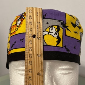 May include: A Halloween-themed scrub cap with a black brim. The cap features a patchwork design with yellow and purple panels. Each panel has a cartoon graphic of a ghost, bats, and a cat. The cap measures approximately 3 inches in height.