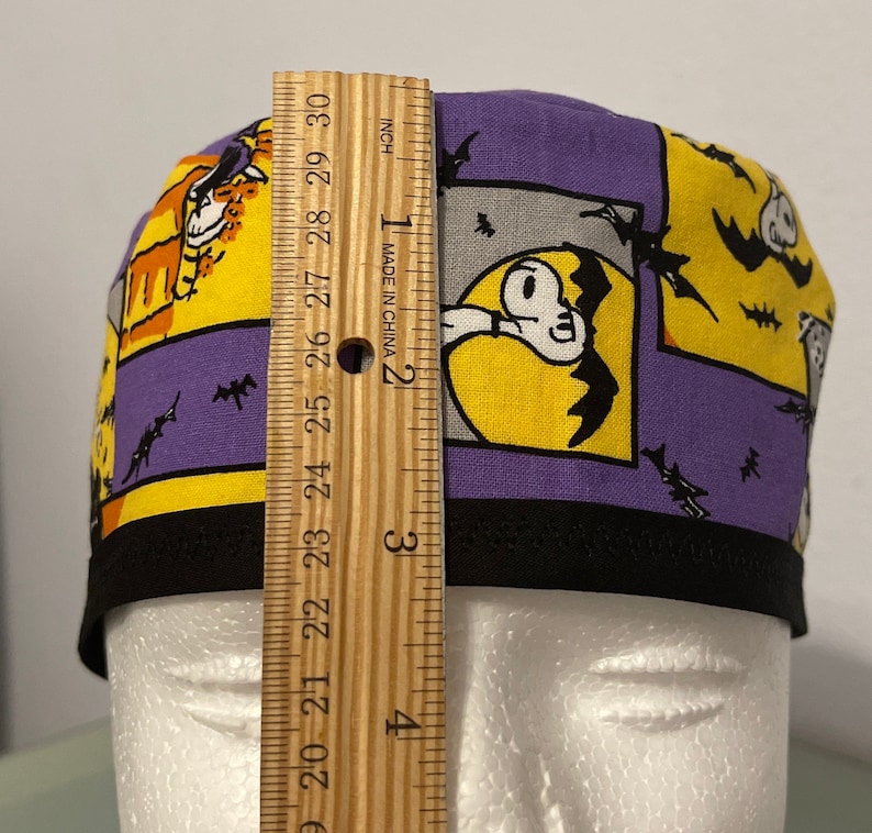 May include: A Halloween-themed scrub cap with a black brim. The cap features a patchwork design with yellow and purple panels. Each panel has a cartoon graphic of a ghost, bats, and a cat. The cap measures approximately 3 inches in height.