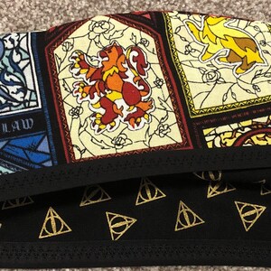 Puede incluir: Un gorro quirúrgico con un patrón de vidrieras de colores con escudos, leones y el texto Ravenclaw, contrastado con una banda de tela negra y una sección inferior decorada con un patrón repetitivo de símbolos triangulares dorados.