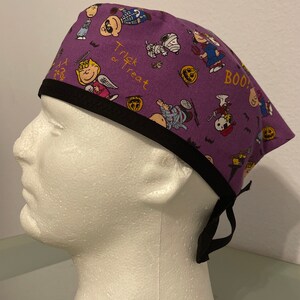 May include: A purple surgical scrub cap featuring a Halloween-themed pattern with cartoon characters, pumpkins, and the words "Trick or Treat" and "BOO!". The cap has a black trim and ties.