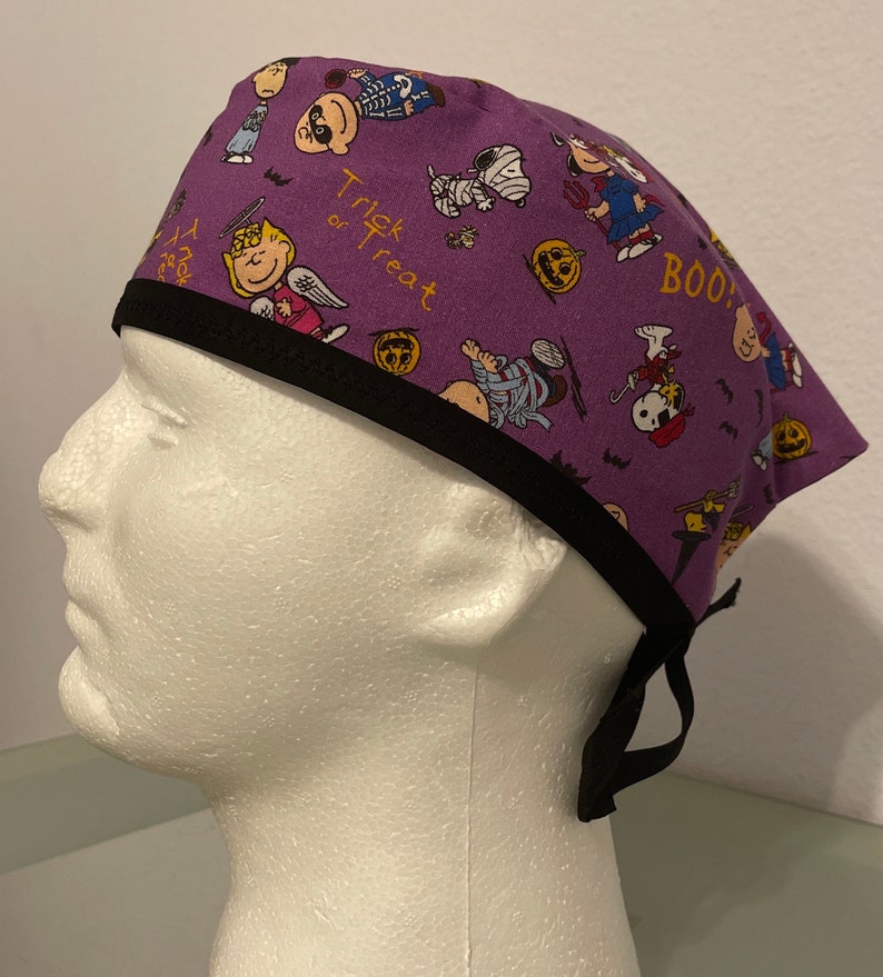 May include: A purple surgical scrub cap featuring a Halloween-themed pattern with cartoon characters, pumpkins, and the words "Trick or Treat" and "BOO!". The cap has a black trim and ties.