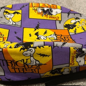May include: A Halloween-themed fabric hat featuring Snoopy in various poses against a backdrop of purple, orange, and yellow squares. The design includes bats, moons, and the words "Trick or Treat". The hat has a black brim and strap.