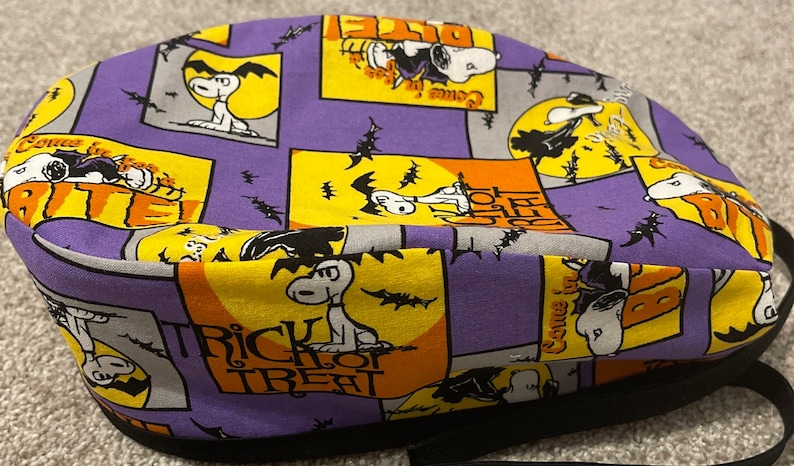 May include: A Halloween-themed fabric hat featuring Snoopy in various poses against a backdrop of purple, orange, and yellow squares. The design includes bats, moons, and the words "Trick or Treat". The hat has a black brim and strap.