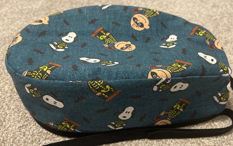 May include: A blue fabric cap featuring a repeating pattern of Snoopy and Charlie Brown dressed as skeletons, with black bat silhouettes. The cap has a black elastic band and is likely for medical or culinary use.