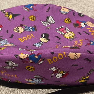 May include: A purple Halloween-themed scrub cap featuring cartoon characters in costumes, pumpkins, bats, and the words "BOO!" and "Trick or Treat". The cap has a black band.