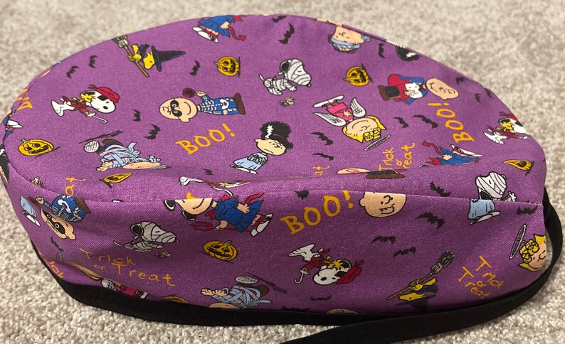 May include: A purple Halloween-themed scrub cap featuring cartoon characters in costumes, pumpkins, bats, and the words "BOO!" and "Trick or Treat". The cap has a black band.