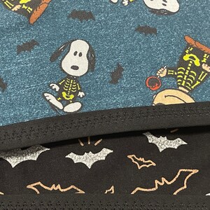 May include: Close-up of two fabric patterns. The top fabric is teal with Snoopy characters in skeleton costumes and black bats. The bottom fabric is black with silver and gold bats.