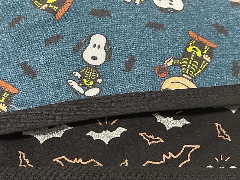 May include: Close-up of two fabric patterns. The top fabric is teal with Snoopy characters in skeleton costumes and black bats. The bottom fabric is black with silver and gold bats.