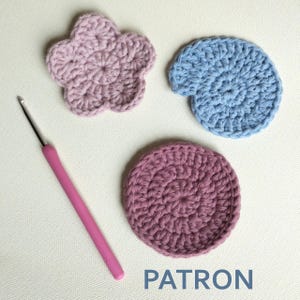 May include: Three crocheted coasters in different shapes: a flower, a shell, and a circle. The coasters are in shades of pink and blue. A pink crochet hook is also in the image.