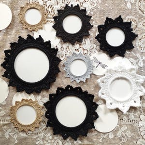 May include: A collection of handmade, starburst-shaped picture frames in various colors, including black, gold, silver, and white. The frames are made of crocheted yarn and have a circular opening for photos or artwork. The frames are arranged on a lace surface.
