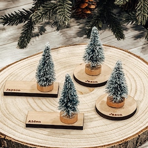 May include: Four miniature Christmas tree place card holders on a wooden slice. Each tree is attached to a wooden base. Two rectangular nameplates read "Jayson" and "Aiden". Two circular nameplates read "Jason". The trees are green with white tips.