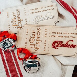 May include: Two wooden Polar Express tickets with the word "Believe" and two silver bells with red ribbons. The tickets have the text "The Bell Still Rings For Those Who Truly Believe". The bells also have the word "Believe".