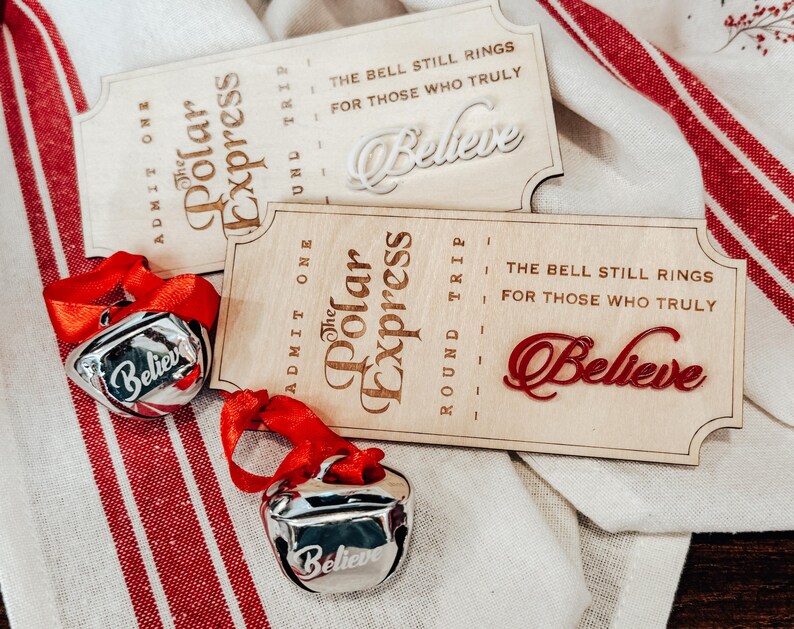 May include: Two wooden Polar Express tickets with the word "Believe" and two silver bells with red ribbons. The tickets have the text "The Bell Still Rings For Those Who Truly Believe". The bells also have the word "Believe".