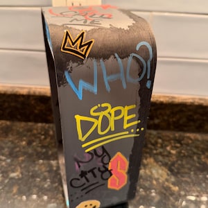 Pode incluir: Um suporte de exibi&ccedil;&atilde;o curvo cinza-escuro com texto e gr&aacute;ficos em estilo grafite. A superf&iacute;cie exibe as palavras CALL ME, WHO?, DOPE e NY CITY, juntamente com uma coroa amarela, uma letra S rosa e um &iacute;cone de rosto sorridente.