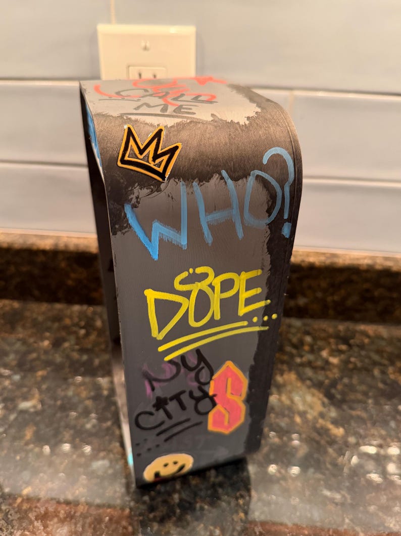 Pode incluir: Um suporte de exibi&ccedil;&atilde;o curvo cinza-escuro com texto e gr&aacute;ficos em estilo grafite. A superf&iacute;cie exibe as palavras CALL ME, WHO?, DOPE e NY CITY, juntamente com uma coroa amarela, uma letra S rosa e um &iacute;cone de rosto sorridente.