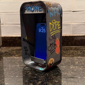 Pode incluir: Um suporte de telefone retangular cinza-escuro em forma de cabine telef&ocirc;nica em miniatura, com textos em estilo grafite como "PHONE", "WHO", "DOPE" e "NY CITY", um &iacute;cone de rosto sorridente amarelo e um smartphone na prateleira interna.