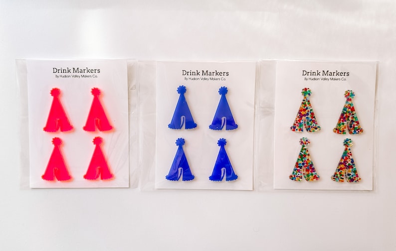 May include: Set of six drink markers featuring party hat designs. The markers are made of acrylic and come in three colors: pink, blue, and multi-colored confetti. Each marker has a small pom-pom on top. The text "Drink Markers" and "By Hudson Valley Makers Co." is printed on each card.