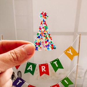 May include: A colorful party tent cake topper with a confetti design. The topper is made of clear plastic and has a white string attached to it. The topper is sitting on a table with a white tablecloth. The letters "PARTY" are spelled out in colorful flags behind the topper.