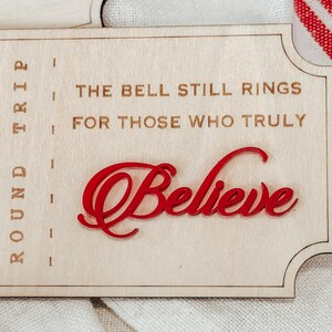 May include: Wooden sign with the text "The Bell Still Rings For Those Who Truly Believe" in a decorative font. The word "Believe" is in red, with a cursive font. The sign is light brown.
