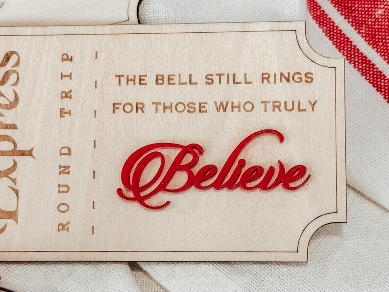 May include: Wooden sign with the text "The Bell Still Rings For Those Who Truly Believe" in a decorative font. The word "Believe" is in red, with a cursive font. The sign is light brown.