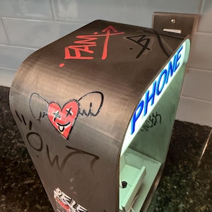 May include: A curved, dark-toned charging station featuring a red heart graphic with wings and X-shaped eyes, the word PHONE in blue lettering, and various graffiti-style markings including the text FAM and RELEASE on its exterior surface.