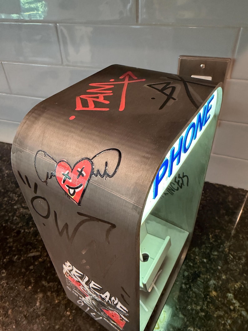 May include: A curved, dark-toned charging station featuring a red heart graphic with wings and X-shaped eyes, the word PHONE in blue lettering, and various graffiti-style markings including the text FAM and RELEASE on its exterior surface.