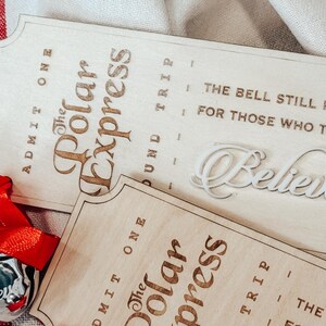 May include: Wooden Polar Express tickets with engraved text and a silver bell ornament with a red ribbon. The tickets read "Admit One" and "The Polar Express" with the phrase "The bell still rings for those who truly believe."