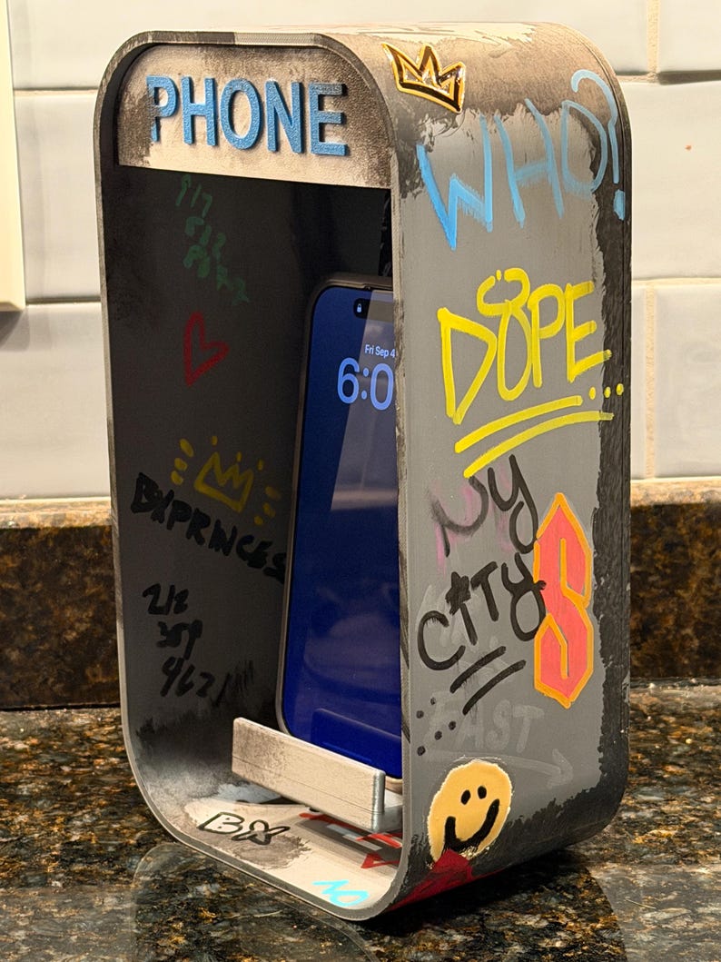 Pode incluir: Um suporte de telefone cinzento e retangular em forma de cabine telef&oacute;nica em miniatura, com grafites como "PHONE", "WHO?", "DOPE" e "NY CITY", uma cara sorridente amarela, uma coroa e um smartphone pousado na prateleira interna.